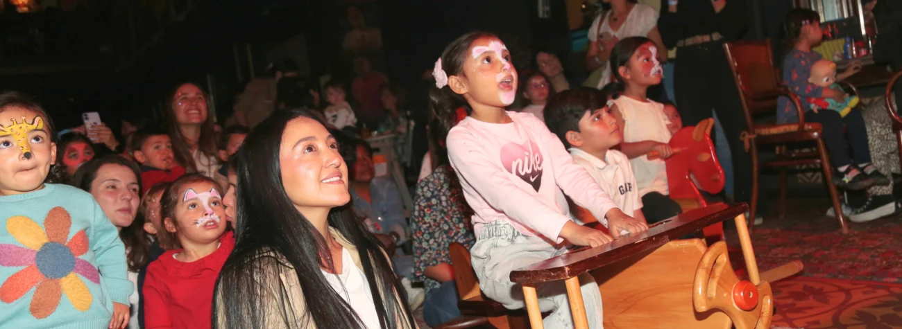 Público infantil y adulto reunido en un espacio cultural para un evento familiar; varios niños interactúan con un juguete con forma de caballito de madera mientras el público observa en un ambiente iluminado y acogedor.
