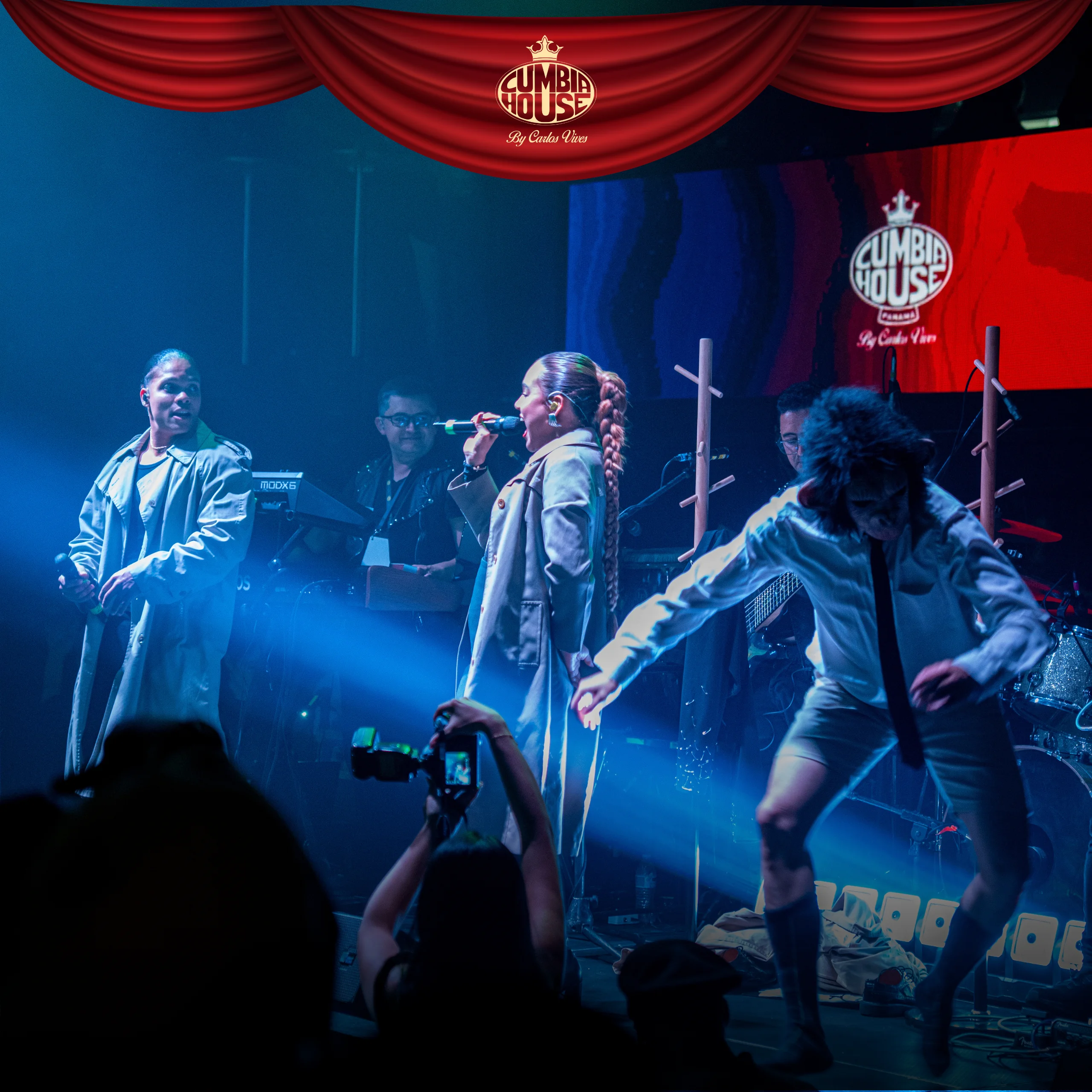 Escenario de concierto en interior con iluminación azul y roja. Varias personas se presentan en vivo: una persona en el centro canta con un micrófono, otra persona a la izquierda sostiene un micrófono y mira hacia el centro, mientras al fondo se observa un teclado y otro integrante del grupo. A la derecha, una persona realiza movimientos de baile frente a una guitarra eléctrica. En la parte superior cuelga un telón rojo con el logotipo “Cumbia House By Carlos Vives”. En primer plano, se ven manos del público levantando teléfonos para grabar la presentación.