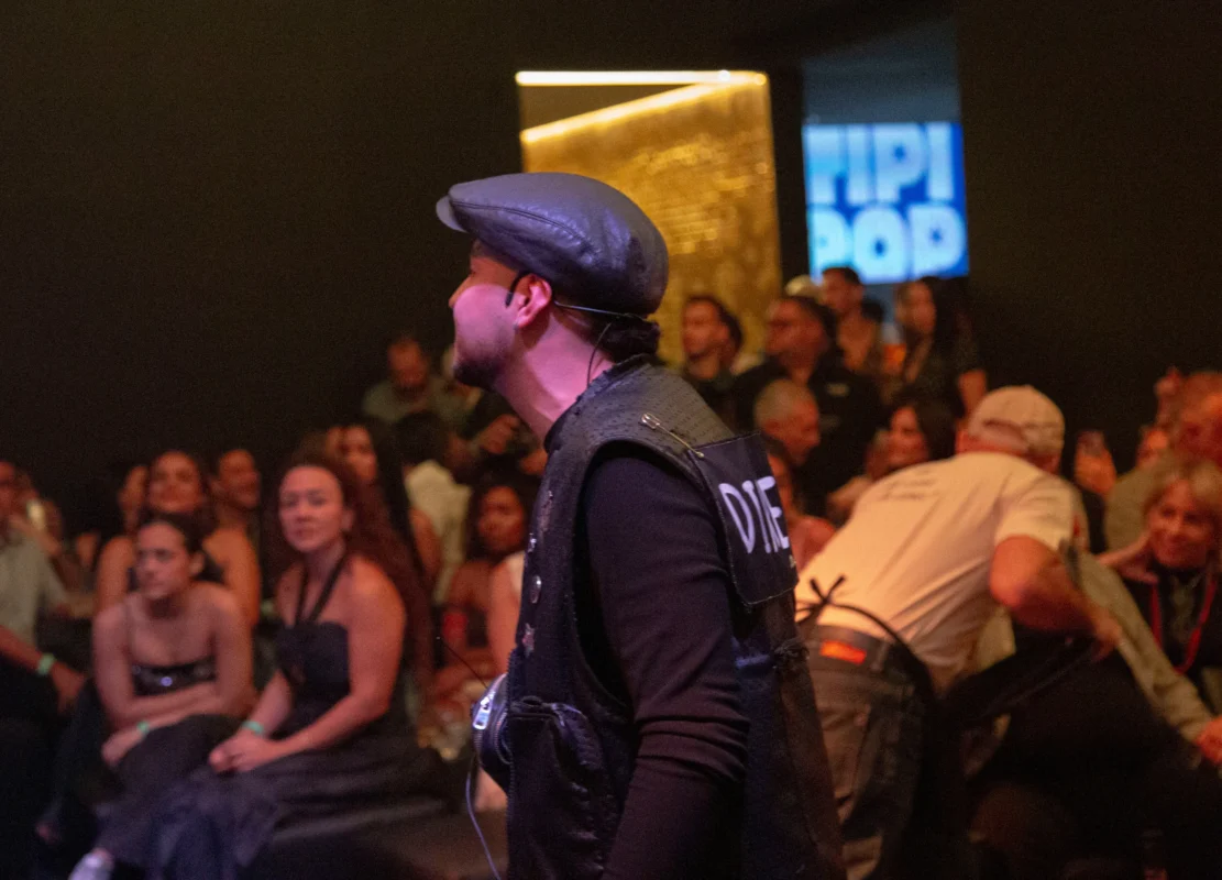 Foto tomada desde el escenario durante un evento en interior. En primer plano, una persona lleva una gorra plana y un chaleco oscuro con letras visibles en la espalda, mirando hacia el público. A la derecha, otra persona se inclina sobre una mesa o equipo técnico. Al fondo se observa una audiencia numerosa sentada y de pie, distribuida en varias filas en un espacio tipo sala de espectáculos. La iluminación es cálida con tonos amarillos y naranjas. En la pared del fondo se aprecia una pantalla o letrero iluminado con el texto “HIPI POD”.