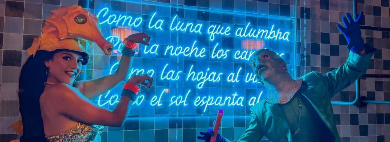 Fotografía tomada en un espacio interior con paredes recubiertas de azulejos oscuros y un letrero de neón azul al fondo. Dos personas posan frente al letrero usando disfraces inspirados en criaturas marinas. A la izquierda, una persona lleva un tocado con forma de caballito de mar naranja y un vestuario brillante; levanta ambos brazos en una pose dinámica. A la derecha, otra persona viste una máscara de pez, un saco verde azulado y guantes azules, sosteniendo un objeto tubular de color naranja. La iluminación azul del neón baña toda la escena y crea un ambiente teatral y festivo.