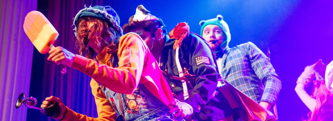 Actuación teatral o musical sobre un escenario iluminado con luces violetas, azules y rosadas. En primer plano aparecen varias personas con vestuarios llamativos y accesorios de fantasía. Una persona sostiene un objeto con forma de paleta helada gigante en una mano y una bocina manual en la otra. Otra persona viste un uniforme decorativo oscuro con hombreras y correas blancas. Detrás, otra persona lleva un traje de cuadros azules y un gorro verde con ojos salientes en la parte superior. Todas las personas usan micrófonos de diadema. Al fondo se observan cortinas oscuras de teatro y, en el extremo derecho, parte de otra persona con un disfraz de color claro. La escena transmite una presentación escénica dinámica y colorida.
